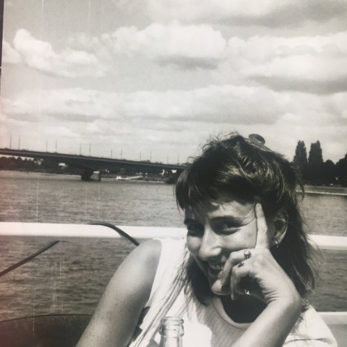 Eine weibliche Person sitzt vor einem Geländer, hinter dem ein Fluss zu sehen ist und lächelt in die Kamera. Das Bild ist schwarz-weiß.