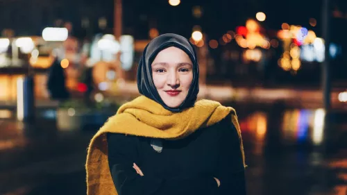 Kübra steht nachts mit einem leuchtend gelben Schal im Freien. Hinter ihr sind viele Lichter zu erkennen.