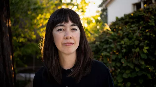 Jenny Odell trägt ein schwarzes Shirt und dunkle Haare. Im Hintergrund sind Büsche, Bäume und ein Haus zu erkennen.