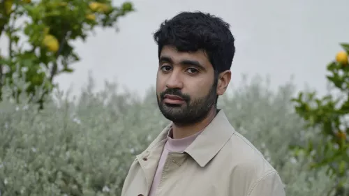 Mohamed trägt einen hellen Trenchcoat. Er steht vor einigen Bäumen und Büschen.