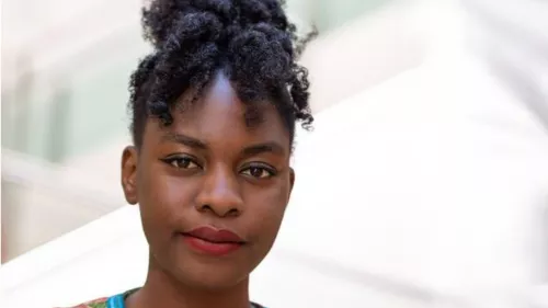 Foto von Esther vor hellem Hintergrund. Sie trägt ihre lockigen Haare hoch zusammengebunden und steht vor einem hellen Hintergrund.