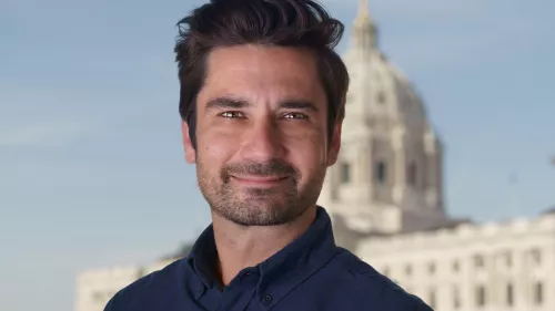 Casey Mock trägt kurze Haare und ein Hemd. Er schaut direkt in die Kamera. Der Hintergrund ist verschwommen und zeigt einen klaren Himmel und einem großen Gebäude. 