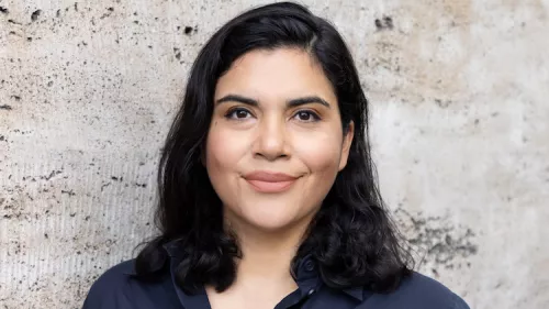 Ein Porträt von Katja Muñoz. Sie trägt eine dunkle Bluse, lehnt an der Wand und lächelt in die Kamera