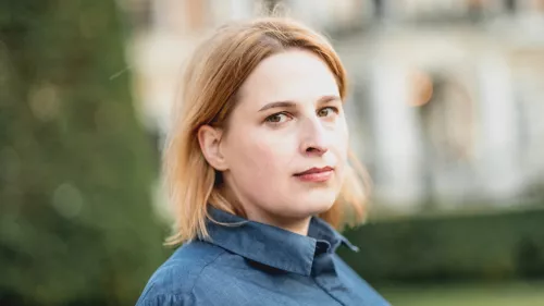 Natascha Strobl schaut seitlich in die Kamera. Im Hintergrund ist ein Gebäude und Baum sowie eine Wiese zu sehen. Natascha Strobl trägt eine Bluse und schulterlanges Haar