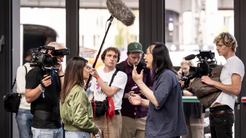 Das Foto zeigt zwei Personen in einem Interview. Außenrum sind Kameras und Mikrofone zu sehen. Diese sind auf die zwei Personen gerichtet. Sie alle stehen vor einer Glasfront.
