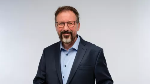 Foto von André Frank Zimpel vor grauem Hintergrund. Er trägt einen Anzug, Brille, Bart und schaut in die Kamera. 