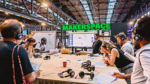 Das Foto zeigt den Makerspace der re:publica. Viele Menschen versammeln sich rund um einee große Holzfläche. Viele von ihnen tragen Kopfhörer. Zettel und Kopfhörer liegen auch auf dem Tisch ab. 