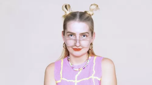 Foto von Isabelle Rogat. Sie trägt zwei Spacebuns, Lippenstift und eine Sonnebrille. Dazu ein Top mit Kette.