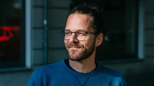 Foto von Arne Semsrott. Er wird von der Sonne angestrahlt, schaut zur Seite. Außerdem trägt er seine Haare im Dutt und eine Brille.