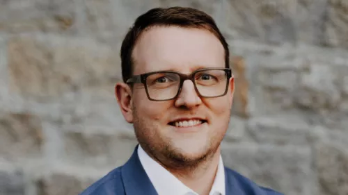 Foto von Matthias Quent. Er steht vor eine Steinwand und lächelt in die Kamera. Dazu trägt er kurze Haare, Anzug und Brille.