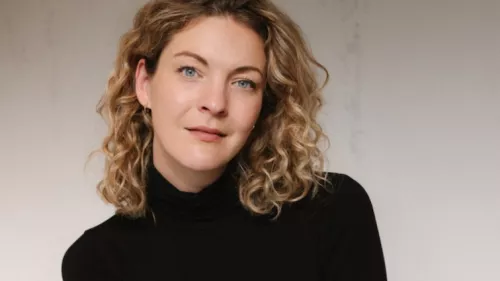 Foto von Sally Lisa Starken. Sie trägt mittellanges lockiges Haar und einen Rollkragen Pullover. Sie sitzt vor einer Betonwand und schaut direkt in die Kamera.
