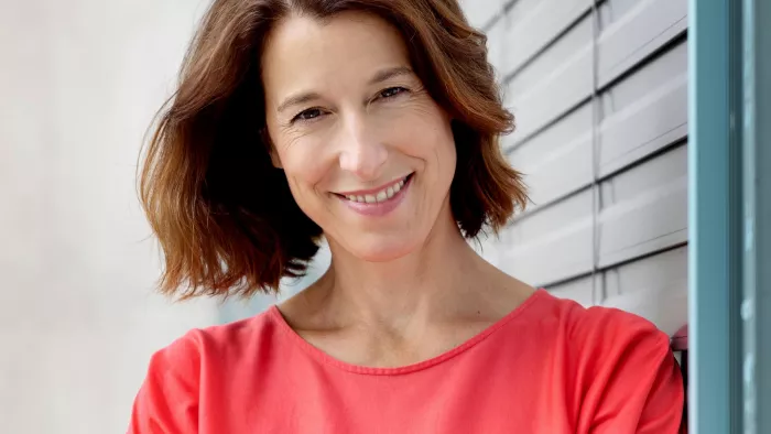 Ellen trägt eine korallenfarbene Bluse und steht vor einer Jalousie. Sie hat schulterlange Haare und lächelt in die Kamera.