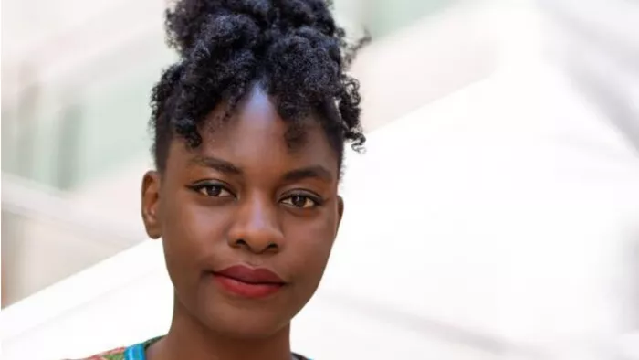 Foto von Esther vor hellem Hintergrund. Sie trägt ihre lockigen Haare hoch zusammengebunden und steht vor einem hellen Hintergrund.
