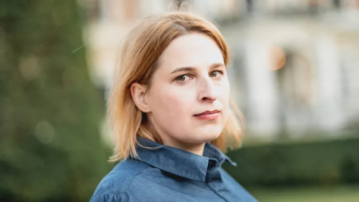 Natascha Strobl schaut seitlich in die Kamera. Im Hintergrund ist ein Gebäude und Baum sowie eine Wiese zu sehen. Natascha Strobl trägt eine Bluse und schulterlanges Haar