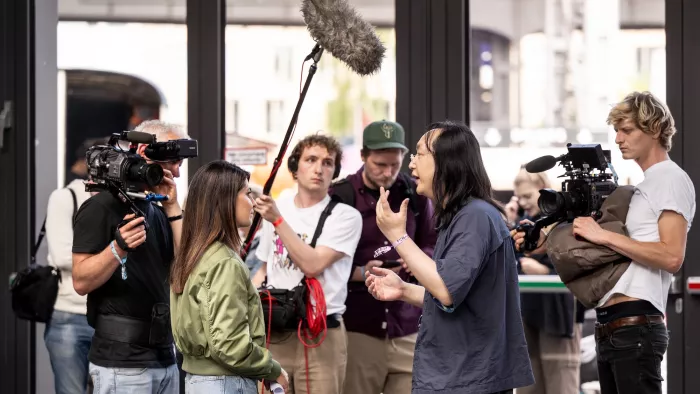 Das Foto zeigt zwei Personen in einem Interview. Außenrum sind Kameras und Mikrofone zu sehen. Diese sind auf die zwei Personen gerichtet. Sie alle stehen vor einer Glasfront.