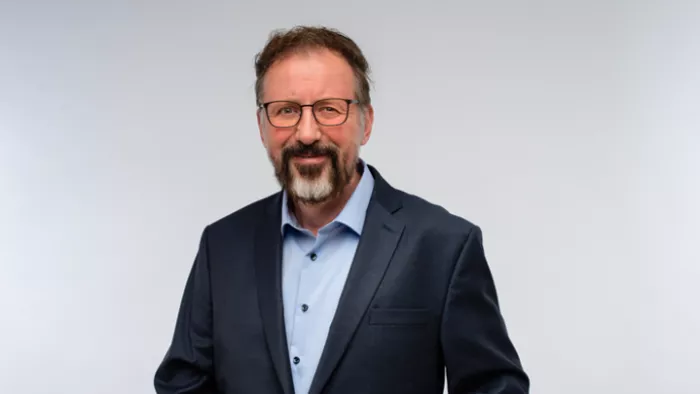 Foto von André Frank Zimpel vor grauem Hintergrund. Er trägt einen Anzug, Brille, Bart und schaut in die Kamera. 