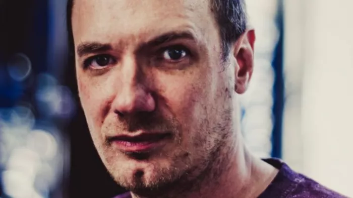 Portraitfoto von Christian Schiffer. Er trägt ein T-Shirt und kurze Haare. Er schaut skeptisch in die Kamera. Das Foto ist mit starkem Kontrast earbeitet. 
