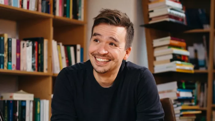 Portraitfoto von Bob Blume in der Natur. Er lächelt seitlich an der Kamera vorbei und trägt einen Pullover. Im Hintergrund ist eine Bücherregalwand zu sehen