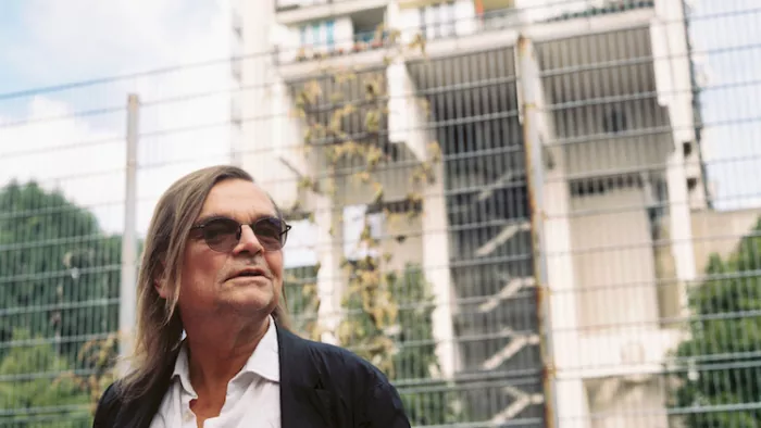 Das Foto zeigt ein Portrait von Diedrich Diederichsen. Er trägt Brille, schulterlanges glattes Haar und schaut seitlich in die Kamera. 