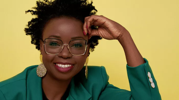 Lunia Hara trägt Ohrringe, Brille, Lippenstift und einen kurzen Afro. Sie stützt ihre Stirn auf ihrer Hand ab und lächelt in die Kamera. Der Hintergrund ist einfarbig  