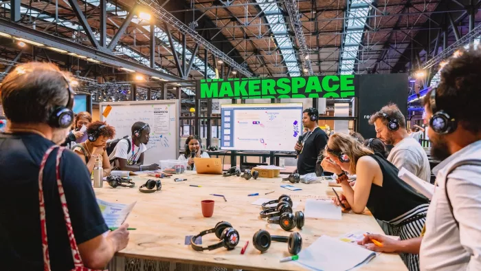 Das Foto zeigt den Makerspace der re:publica. Viele Menschen versammeln sich rund um einee große Holzfläche. Viele von ihnen tragen Kopfhörer. Zettel und Kopfhörer liegen auch auf dem Tisch ab. 