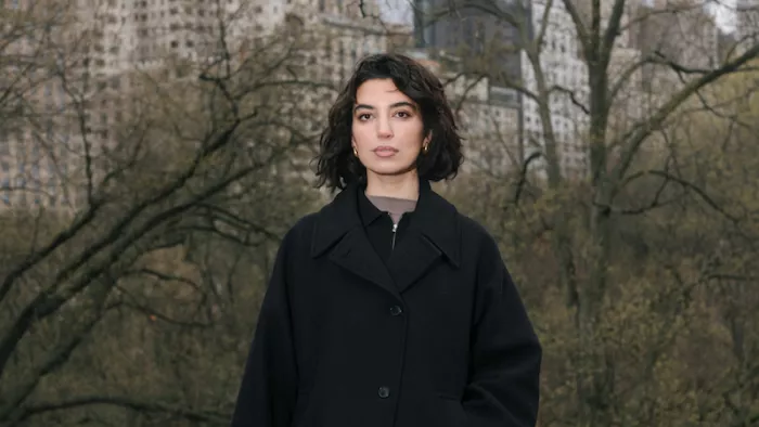 Das Foto zeigt Aya Jaff in New York. Sie steht vor Bäumen und Gebäuden und trägt einen Mantel sowie kinnlanges Haar