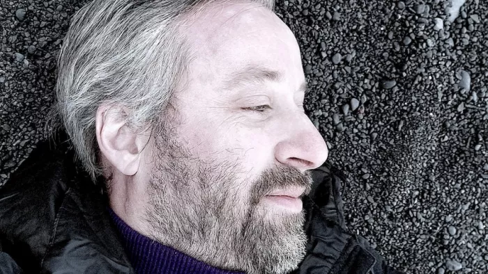 Portrait von Robin Berjon. Er liegt auf dem Boden und schaut nach rechts. Dabei trägt er eine Jacke, Rollkragenpullover sowie kurze Haare und Bart.