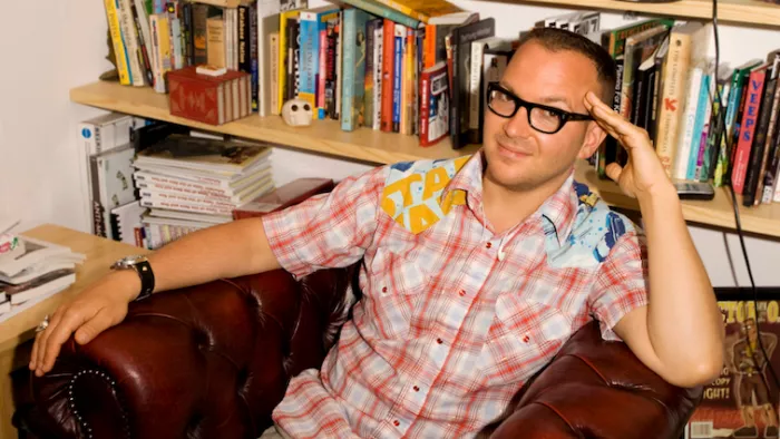 Foto von Cory Doctorow. Er sitzt in einem Sessel und stützt einem Arm auf einer Lehne ab. Cory trägt kurze Haare, Brille und einen Pullover