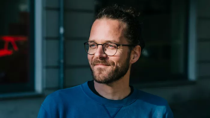 Foto von Arne Semsrott. Er wird von der Sonne angestrahlt, schaut zur Seite. Außerdem trägt er seine Haare im Dutt und eine Brille.