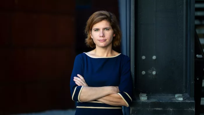 Foto von Ingrid Brodnig. Sie lehnt sich an eine Wand an und hält ihre Arme verschränkt. Sie schaut direkt in die Kamera