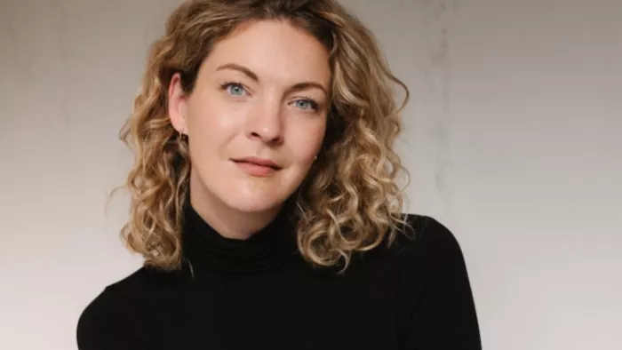 Foto von Sally Lisa Starken. Sie trägt mittellanges lockiges Haar und einen Rollkragen Pullover. Sie sitzt vor einer Betonwand und schaut direkt in die Kamera.