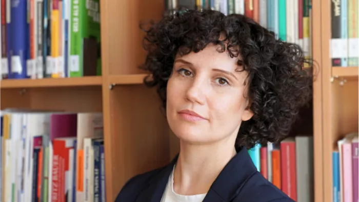 Foto von Siri Hummel. Sie trägt kurze lockige Haare und steht vor einer Bücherwand. Sie schaut direkt in die Kamera