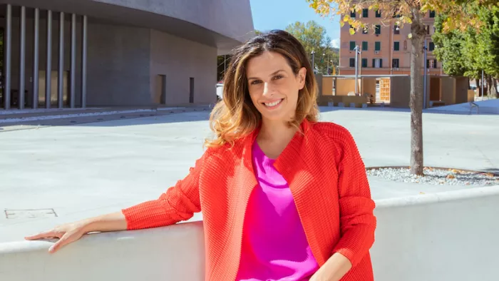 Foto von Francesca Bria. Sie sitzt auf einer Beton-Bank vor einem Gebäude. Eine Hand liegt auf der Lehne auf. Sie trägt ein Langarmshirt und Jacke.