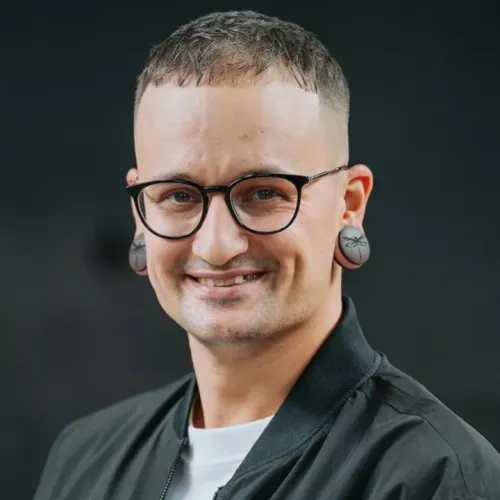 Porträtfoto von Adrian Rosetta. Er trägt eine Brille, kurze Haare und lächelt leicht. Dunkle Kleidung, neutraler Hintergrund.