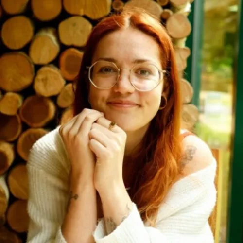 Headshot of Izzie, a person with red hair and glasses, smiling with hands clasped, seated near a wall of stacked wood.