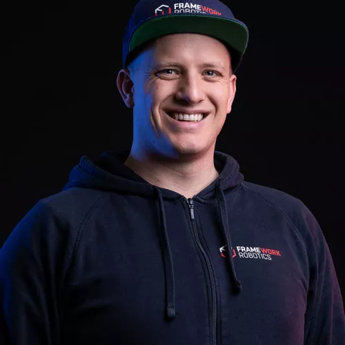 Porträt eines lächelnden Mannes vor schwarzem Hintergrund, der einen dunkelblauen Hoodie und eine Kappe mit dem Logo von „FRAMEWORK ROBOTICS“ trägt.