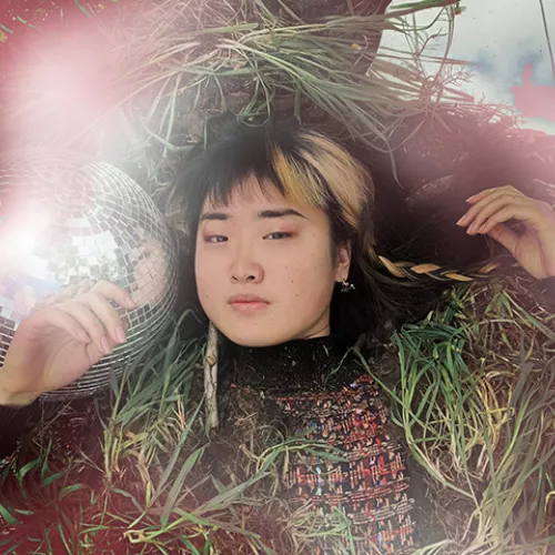 Alice holds a knowing expression as she lies on a bed of soil, surrounded by grass, reflective surfaces, and mystical red-white air