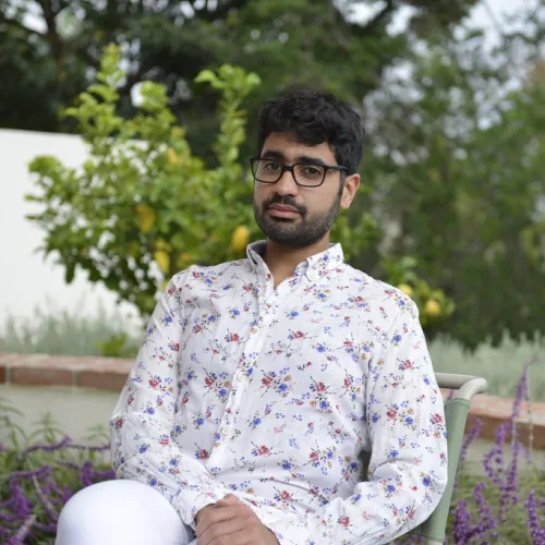 Auf einem Gartenstuhl sitzt Mohamed Amjahid mit einer weißen Hose und einem weißen geblümten Hemd. Er trägt eine Brille und blickt direkt in die Kamera. Hinter ihm sind Bäume und Pflanzen. 