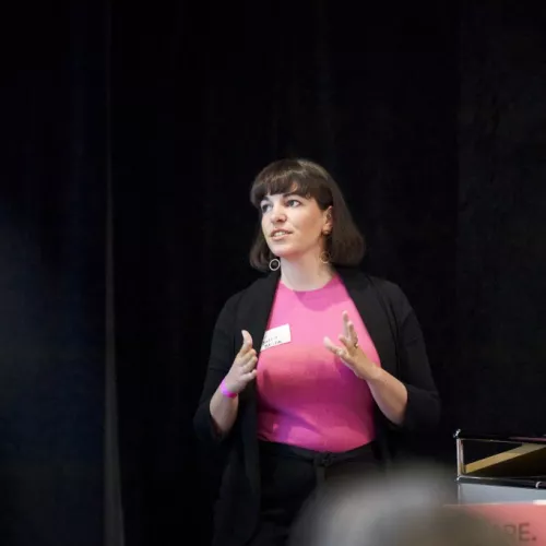 A white woman with brown hair, who is wearing a pink shirt and a black jacket, is speaking to an audience. She looks to one side. Her hands are in action, as she speaks. 