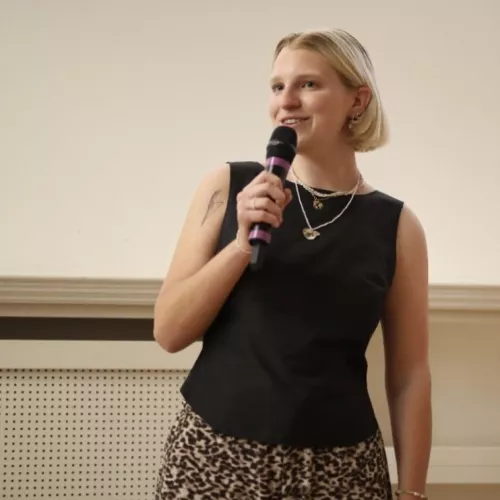Eine Person spricht lächelnd in ein Mikro. Sie trägt ein schwarzes Oberteil und hat kinnlange, blonde Haare.