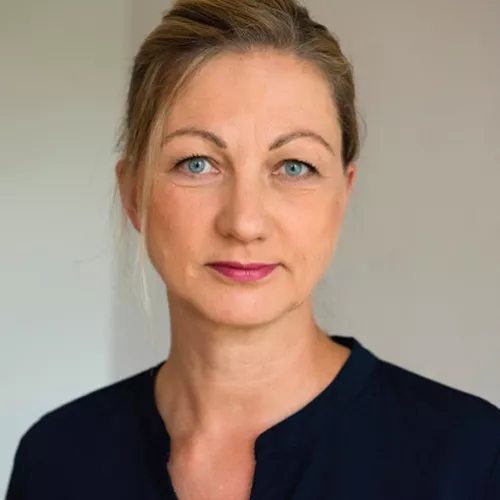 Man sieht eine Frau, die direkt in die Kamera blickt und leicht verschmitzt lächelt. Sie hat eine schwarze Bluse an, ihre blonden Haare sind zurück gesteckt, sie hat blaue Augen.