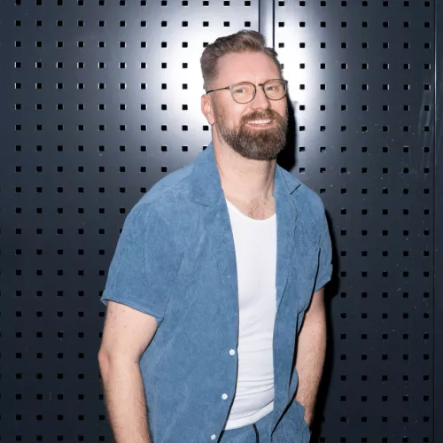 Moderator Mathias Furch steht vor einer dunklen Metallwand. Er trägt ein blaues offenes Hemd, darunter ein weißen T-Shirt. Mathias lacht herzlich auf dem Bild und schaut direkt in die Kamera.