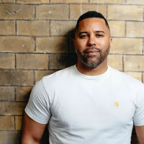 The picture shows a light skinned black man, with close cropped hair and beard. He has broad shoulders and wears a white T-Shirt. He stands infront of a wall. 