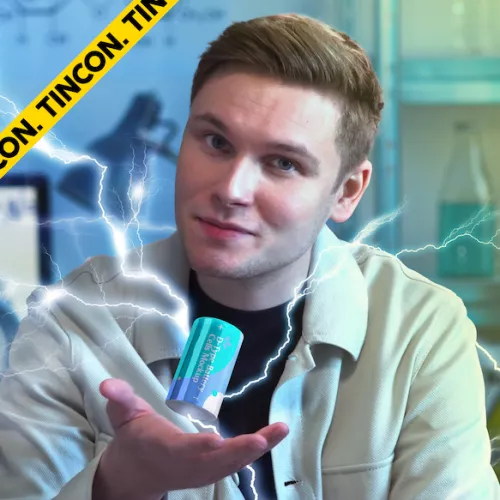 DoktorWissenschaft hält eine blaue Batterie vor die Kamera aus der weiße Blitze kommen. Er trägt einen weißen Kittel und darunter ein schwarzes Oberteil. Es ist nur sein Oberkörper zu sehen, er hat kurze blonde Haare und guckt mit schrägem Kopf in die Kamera