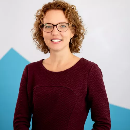 Das Bild zeigt eine freundliche, lächelnde Frau mit kurzen, lockigen, blond-rötlichen Haaren und einer Brille. Sie trägt ein weinrotes, strukturiertes Oberteil mit dreiviertellangen Ärmeln. Im Hintergrund ist eine helle Wand mit einem blauen, diagonal verlaufenden Akzent zu sehen. Die Frau steht aufrecht und wirkt professionell und sympathisch.
