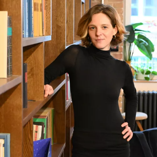 Theresa Schuhmann steht neben einem hohen Holzbücherregal, eine Hand in der Hüfte, die andere am Regal. Sie trägt ein schwarzes Rollkragenoberteil und lächelt leicht in die Kamera.