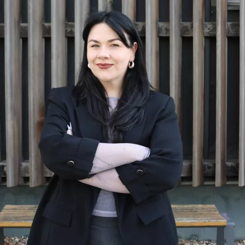 Das Foto zeigt Katharina Rein. Sie steht vor einer holzvertäfelten Wand. Ihre Arme sind verschränkt, sie lächelt und trägt einen schwarzen Blazer.