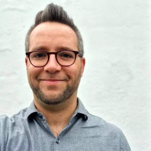 Mann im grauen Hemd mit Brille, Bart und leichtem Iro-Schnitt vor weißer Mauer 