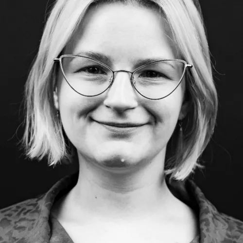 Black and white portrait of Lisa Schmechel. She has a blonde bob and wears cateye-style glasses.