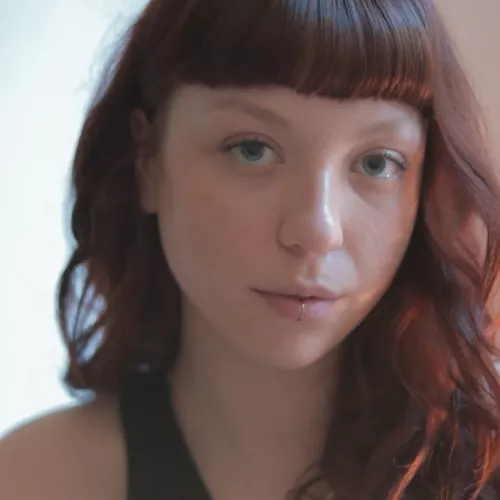 Foto von Regisseurin Karoline Rößler, sie lächelt leicht, hat rote Haare mit einem kurzen Pony und trägt ein schwarzes Top.